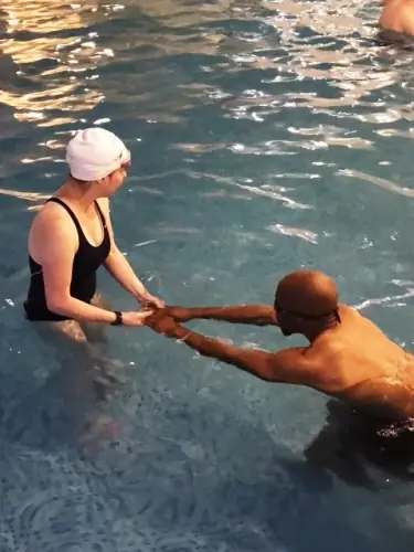 An instructor holding a student's hands as she floats, white cap, teal water
