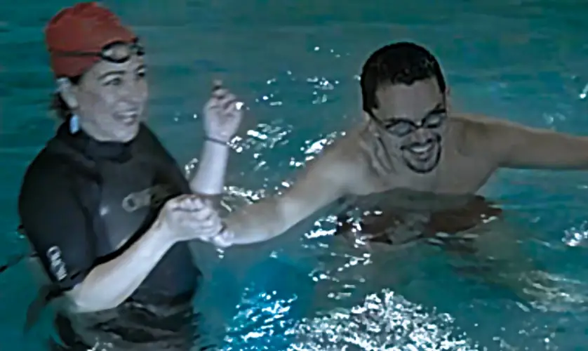 An instructor in a dark wetsuit laughing with an adult student in the pool