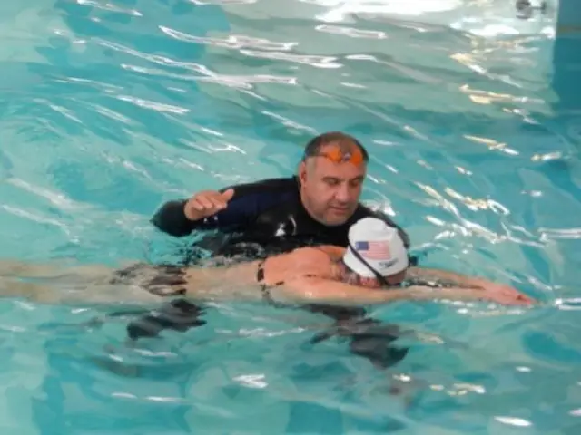 An instructor in a wetsuit supporting a swimmer in a USA cap in bright water