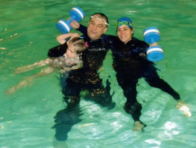Two instructors in wetsuits holding a young swimmer with foam dumbbells
